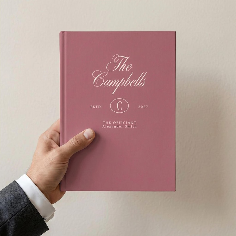 May include: A dusty rose hardcover book featuring white text that reads The Campbells, ESTD 2027, a central letter C inside an oval, and the text THE OFFICIANT Alexander Smith printed on the front cover in a clean, centered layout.
