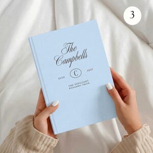 May include: A light blue hardcover book with black text that reads The Campbells, ESTD, 2027, and THE OFFICIANT Alexander Smith, featuring a central oval containing the letter C. The cover has a smooth, matte finish and a clean, centered layout.
