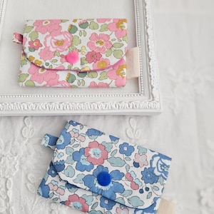 May include: Two small floral print wallets. One is pink with a pink button, the other is blue with a blue button. Both have a small tab and a tag. The wallets are on a white surface.
