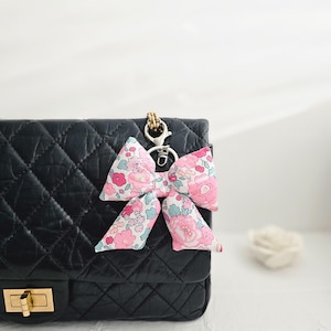 Op de afbeelding: Een bloemenstrik sleutelhanger met roze, blauwe en witte bloemen, bevestigd aan een zwarte gewatteerde handtas. De strik is gemaakt van stof en heeft een goudkleurige sluiting.