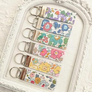 Könnte beinhalten: Eine Auswahl an Schlüsselanhängern aus Stoff mit Blumenmuster und silberfarbenen Metallbeschlägen. Jeder Schlüsselanhänger zeigt ein anderes Blumenmuster in Lila, Blau, Rosa, Gelb und Grün. Die Schlüsselanhänger liegen auf einem weißen Tablett.
