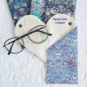 Peut inclure: Un étui à lunettes en tissu en forme de cœur avec un intérieur crème et un motif floral bleu à l'extérieur. L'étui est doté d'une fermeture à pression et du texte Opens into a Heart. Des lunettes à monture noire reposent sur l'étui ouvert.