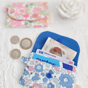 May include: A small, floral-patterned wallet with a blue interior, holding cards and cash. The wallet features a blue snap closure and is made of fabric with blue and pink flowers. A second wallet with a pink floral pattern is in the background.