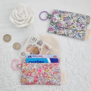 May include: Two small floral print coin purses with a pink and purple color scheme. One purse is open, revealing a UK driving license and an Oyster card. The other purse is closed. Both purses have a ring attached.
