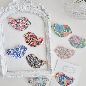 Può includere: Una collezione di fermagli per capelli a forma di uccello in tessuto floreale in vari colori, tra cui rosa, blu e rosso. Alcuni fermagli sono esposti all'interno di una cornice bianca ornata, altri sono disposti su una superficie bianca. È presente anche una carta con un disegno floreale di uccello.
