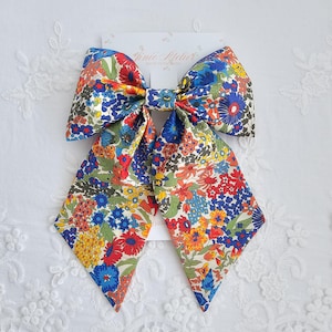 Könnte beinhalten: Eine große Haarschleife aus Stoff mit einem lebendigen Blumenmuster aus roten, blauen, gelben und grünen Blüten auf cremefarbenem Grund. Die Schleife ist auf einer weißen Karte mit dem Aufdruck Jinie Atelier Made in England befestigt.