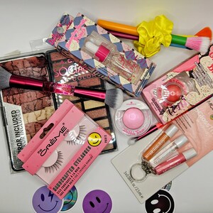 May include: A collection of beauty products including a large eyeshadow palette, a dual-ended makeup brush with a pink handle, a bottle of illuminating primer, a cream blush, a pair of false eyelashes, a pink powder compact, and a lip gloss trio set.