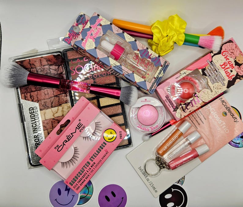 May include: A collection of beauty products including a large eyeshadow palette, a dual-ended makeup brush with a pink handle, a bottle of illuminating primer, a cream blush, a pair of false eyelashes, a pink powder compact, and a lip gloss trio set.