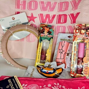 May include: A collection of accessories including a beige pearl-embellished headband, a pink bandana, a cowboy hat-shaped hair clip, and two packages of lip gloss labeled Shine Gloss and Wrangler Pout Pair, all set against a pink Howdy print fabric.