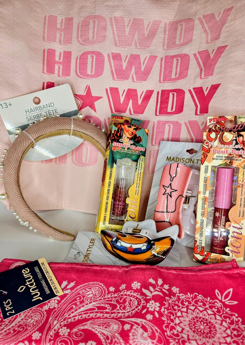 May include: A collection of accessories including a beige pearl-embellished headband, a pink bandana, a cowboy hat-shaped hair clip, and two packages of lip gloss labeled Shine Gloss and Wrangler Pout Pair, all set against a pink Howdy print fabric.
