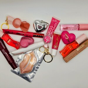 May include: A collection of beauty products including a Fafamoon lip tube, a red lip gloss, a pink heart-shaped compact, a silver heart keychain, an Essence Kiss Bomb tube, a Thalia Sodi lip gloss, a lip oil, and a gold-toned lip-shaped mask.