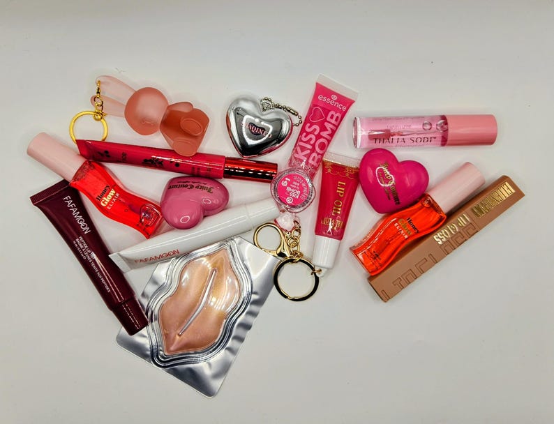 May include: A collection of beauty products including a Fafamoon lip tube, a red lip gloss, a pink heart-shaped compact, a silver heart keychain, an Essence Kiss Bomb tube, a Thalia Sodi lip gloss, a lip oil, and a gold-toned lip-shaped mask.