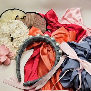 May include: A collection of hair accessories including a grey glittery headband with white pearls, a pink flower-shaped claw clip, various satin bows in red, orange, and light pink, a white lace scrunchie, and ruffled tulle scrunchies in cream and tan.