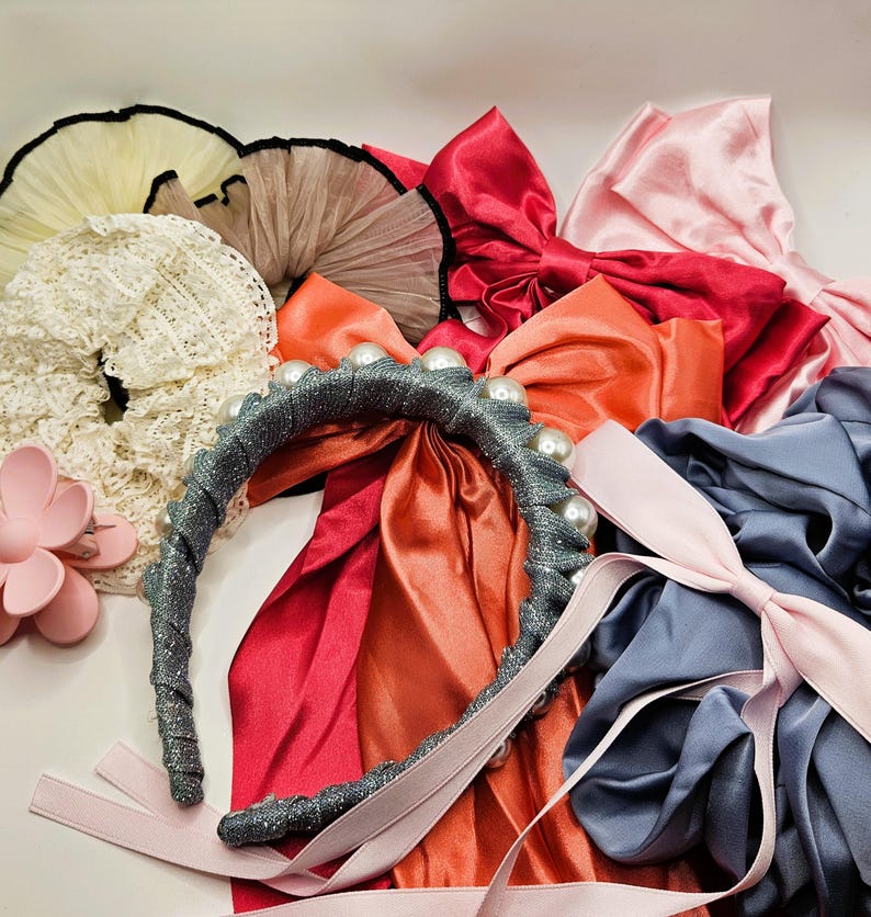 May include: A collection of hair accessories including a grey glittery headband with white pearls, a pink flower-shaped claw clip, various satin bows in red, orange, and light pink, a white lace scrunchie, and ruffled tulle scrunchies in cream and tan.