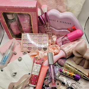 May include: A collection of beauty and self-care items including a Paris Hilton gift set, a heart-shaped eyeshadow palette, makeup brushes with crystal handles, a lip butter balm, lip gloss, a lint roller, a hair clip, and various skincare products.