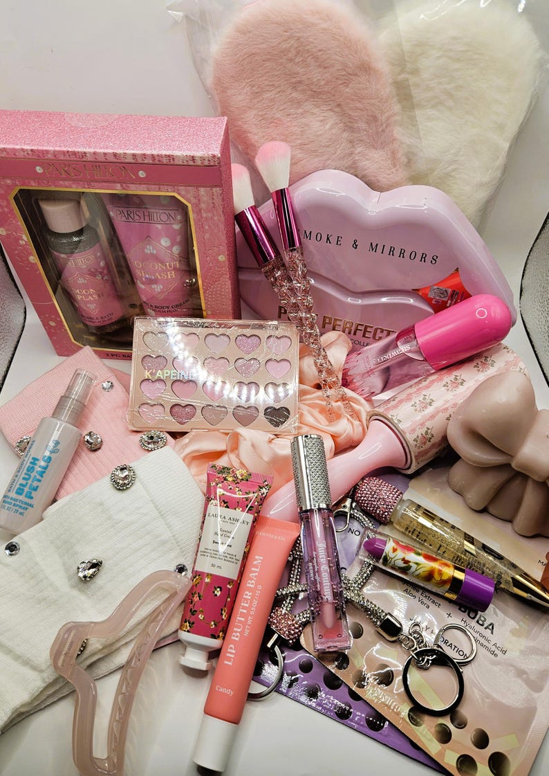 May include: A collection of beauty and self-care items including a Paris Hilton gift set, a heart-shaped eyeshadow palette, makeup brushes with crystal handles, a lip butter balm, lip gloss, a lint roller, a hair clip, and various skincare products.
