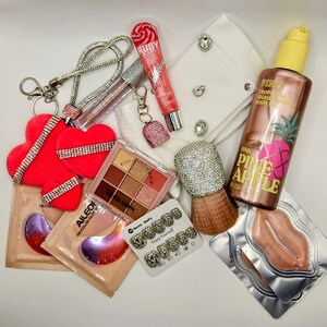 May include: A collection of beauty and fashion items including three red heart-shaped ornaments with rhinestone bands, a tube of Ruby Kisses lip gloss, a Pink Pineapple body oil bottle, a nine-color eyeshadow palette, lip masks, and nail tips.
