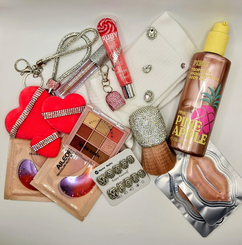 May include: A collection of beauty and fashion items including three red heart-shaped ornaments with rhinestone bands, a tube of Ruby Kisses lip gloss, a Pink Pineapple body oil bottle, a nine-color eyeshadow palette, lip masks, and nail tips.