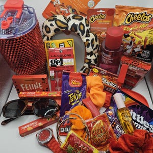 May include: A collection of items including a red studded tumbler, a leopard print headband, Cheetos snacks, Takis, Hot Pops, Skittles, L.A. Colors nail tips, a Pink body mist, sunglasses, a red watch, orange scrunchies, and various beauty products.