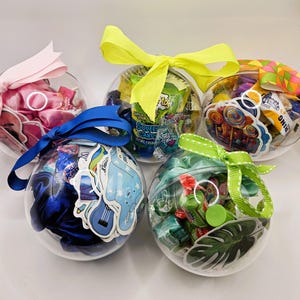 May include: Five clear plastic ornaments filled with various items, including candy, stickers, and hair accessories. Each ornament is tied with a colorful ribbon, such as pink, blue, yellow, and green. The ornaments are arranged on a white surface.