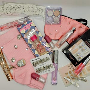 May include: A collection of beauty and fashion items including pink ribbed socks with rhinestones, a Kiss Jewel press-on nail set, a pink sleep mask with pearls, various lip glosses, a primer bottle, a small eyeshadow palette, and hair clips.