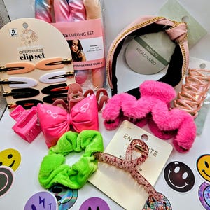May include: A collection of hair accessories including a tie-dye curling set, a pink headband with gold chain detail, a bright pink bow clip, a fuzzy pink claw clip, a neon green scrunchie, a glittery hair clip, and various smiley face stickers.