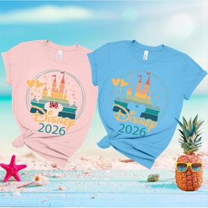 May include: Two t-shirts, one pink and one blue, featuring a Disney castle design. The graphic includes the text "Disney 2026" and a Mickey Mouse head. A pineapple wearing sunglasses and a starfish are in the foreground.