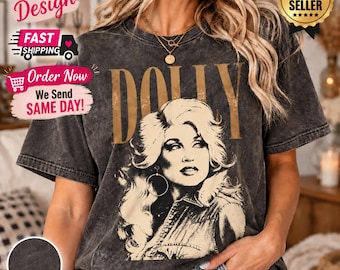 May include: A charcoal grey acid-wash t-shirt featuring a large, tan-colored graphic print of a woman with voluminous hair and the word DOLLY printed in bold, tan capital letters above the portrait. The shirt has short sleeves and a crew neckline.