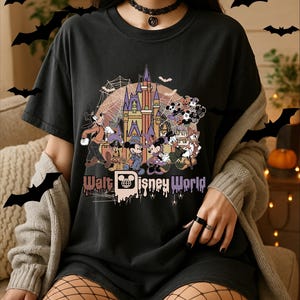May include: A black t-shirt featuring a graphic print of the Walt Disney World castle with Mickey Mouse, Minnie Mouse, Goofy, Donald Duck, and other characters in Halloween costumes, surrounded by spiderwebs and bats against a moon background.
