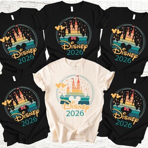 May include: A selection of black and cream-coloured t-shirts with a colourful graphic design. The design features a castle silhouette, Mickey Mouse ears, and the text "Disney 2026" within a circular frame. The shirts are short-sleeved.
