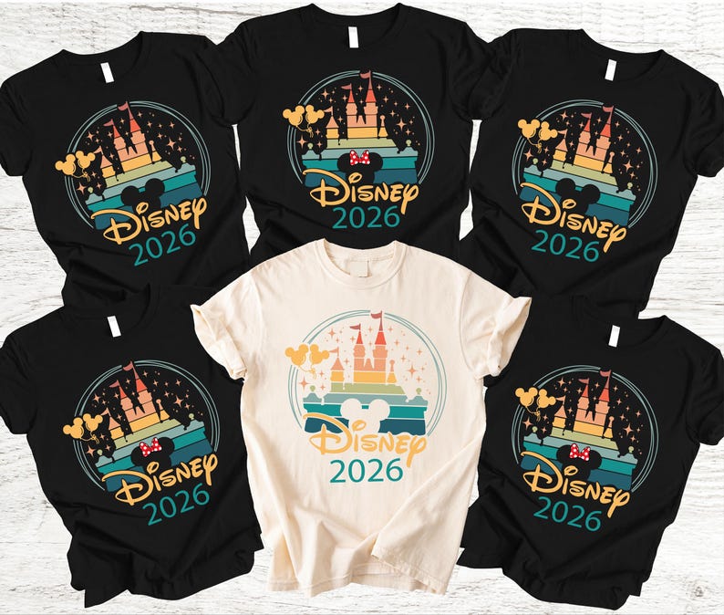 May include: A selection of black and cream-coloured t-shirts with a colourful graphic design. The design features a castle silhouette, Mickey Mouse ears, and the text "Disney 2026" within a circular frame. The shirts are short-sleeved.