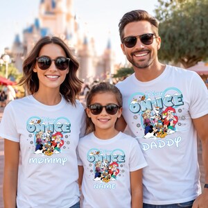 A family of three, consisting of a mother, father, and child, is standing together and smiling. They are all wearing white t-shirts with Disney characters and the text "Disney ONICE".