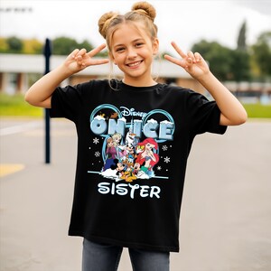 A young girl wearing a black t-shirt with a Disney-themed graphic and text, smiling and making a peace sign gesture.