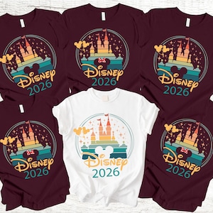 May include: Several maroon and white t-shirts with a colourful graphic design. The design features a castle, Mickey Mouse ears, and the text "Disney 2026" within a circular frame. The shirts are short-sleeved.