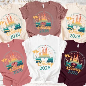 May include: Various coloured t-shirts with a castle design and the text "Disney 2026". The design includes a castle, Mickey Mouse ears, and a rainbow-coloured background. The shirts are available in different colours, including white, pink, and burgundy.
