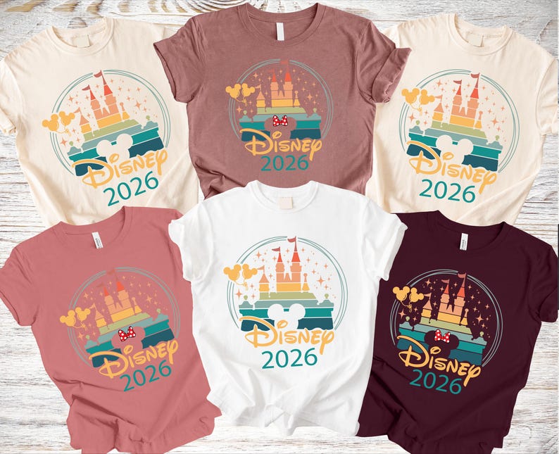 May include: Various coloured t-shirts with a castle design and the text "Disney 2026". The design includes a castle, Mickey Mouse ears, and a rainbow-coloured background. The shirts are available in different colours, including white, pink, and burgundy.