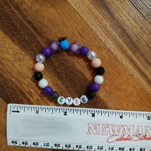 May include: A beaded bracelet with the name "EVIE" spelled out in white letter beads. The bracelet features a mix of colorful beads, including purple, black, white, and peach. A ruler is next to the bracelet.