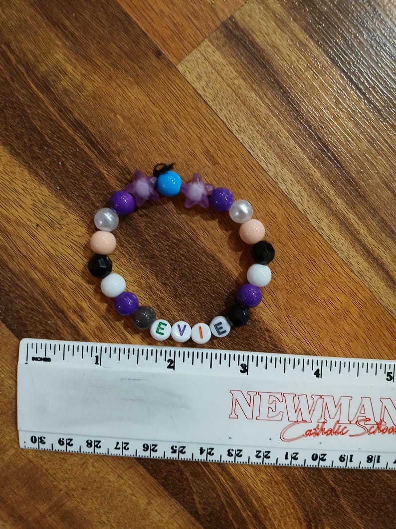 May include: A beaded bracelet with the name "EVIE" spelled out in white letter beads. The bracelet features a mix of colorful beads, including purple, black, white, and peach. A ruler is next to the bracelet.