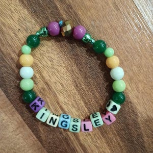 May include: A beaded bracelet with the name "KINGSLEY" spelled out in colorful letter beads. The bracelet features a mix of green, purple, white, and yellow beads. The bracelet is on a wooden surface.