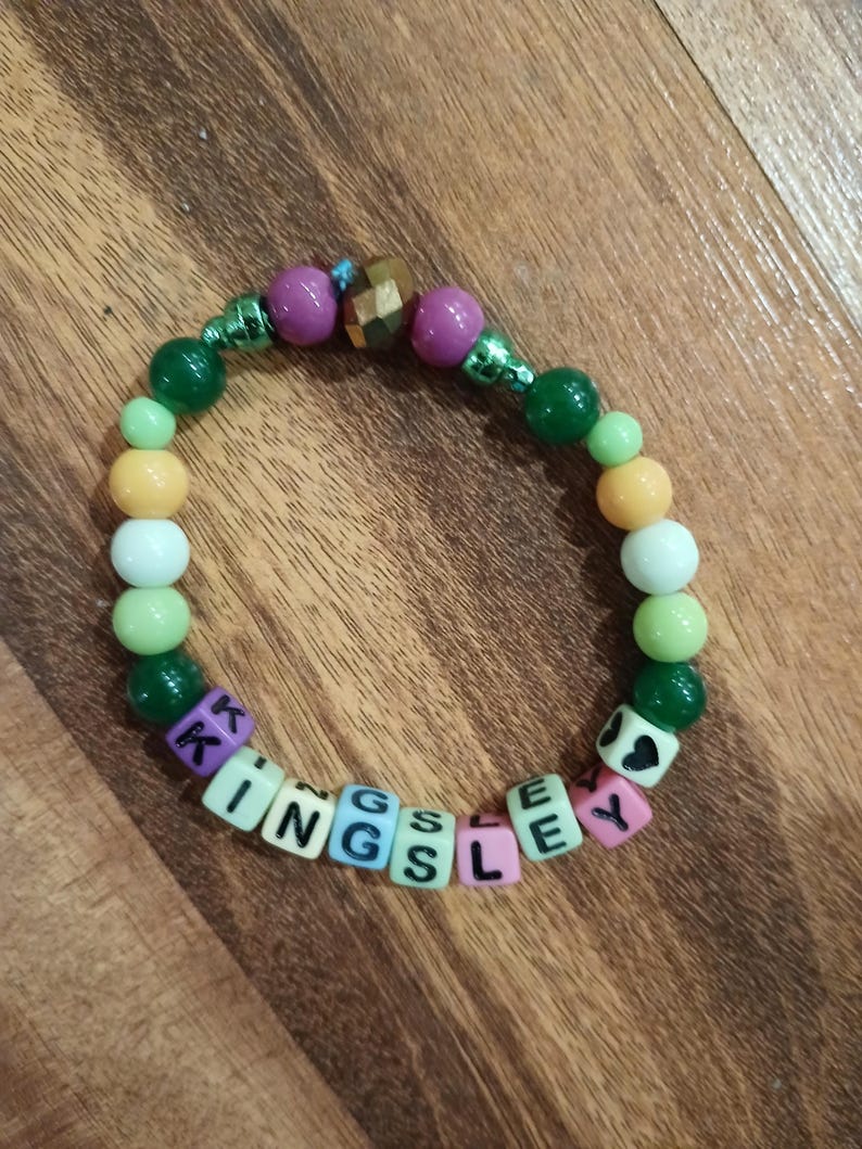May include: A beaded bracelet with the name "KINGSLEY" spelled out in colorful letter beads. The bracelet features a mix of green, purple, white, and yellow beads. The bracelet is on a wooden surface.