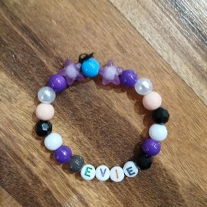 May include: A beaded bracelet with the name "EVIE" spelled out in white letter beads. The bracelet features a variety of beads in colors like purple, black, white, and pink, along with star-shaped and round beads. A small blue bead is also present.