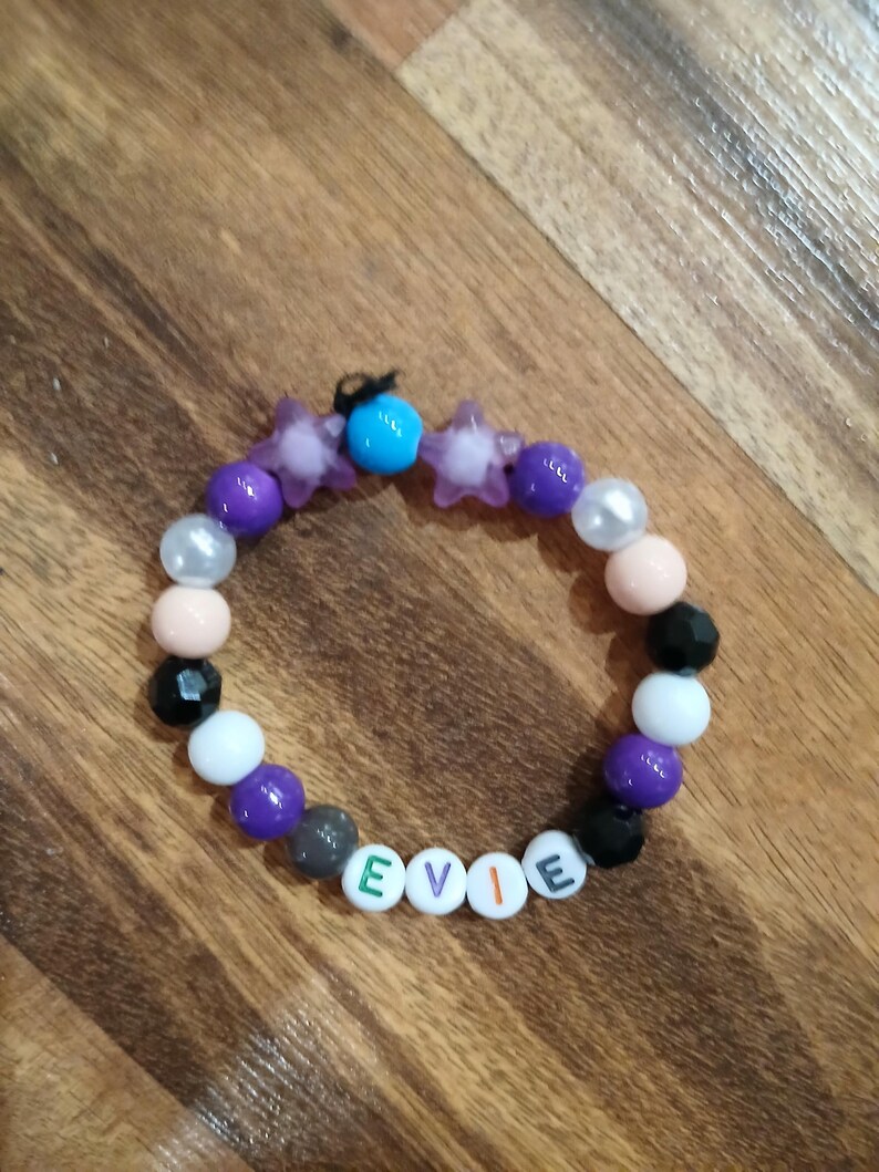 May include: A beaded bracelet with the name "EVIE" spelled out in white letter beads. The bracelet features a variety of beads in colors like purple, black, white, and pink, along with star-shaped and round beads. A small blue bead is also present.