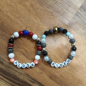 Puede incluir: Dos pulseras de cuentas con los nombres "ALOSA" y "RIDEN" deletreados con cuentas de letras blancas. Las pulseras presentan una mezcla de cuentas coloridas, incluyendo blanco, rojo, negro, gris y dorado.