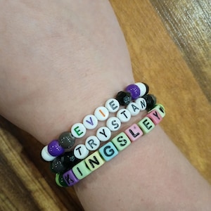 May include: Three beaded bracelets spelling out "EVIE", "TRYSTAN", and "KINGSLEY" in white letter beads. The bracelets feature a mix of colorful beads, including green, purple, black, and white, strung on elastic.