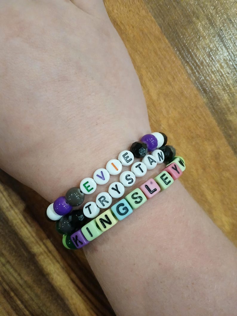 May include: Three beaded bracelets spelling out "EVIE", "TRYSTAN", and "KINGSLEY" in white letter beads. The bracelets feature a mix of colorful beads, including green, purple, black, and white, strung on elastic.