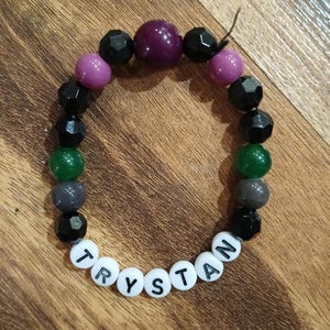 May include: A beaded bracelet with the name "TRYSTAN" spelled out in white letter beads. The bracelet features a mix of black, purple, green, and gray beads. The beads are various shapes and sizes, creating a colorful accessory.