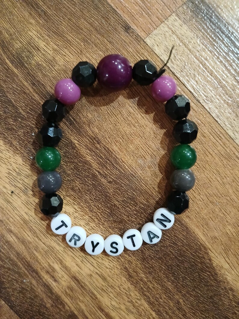 May include: A beaded bracelet with the name "TRYSTAN" spelled out in white letter beads. The bracelet features a mix of black, purple, green, and gray beads. The beads are various shapes and sizes, creating a colorful accessory.