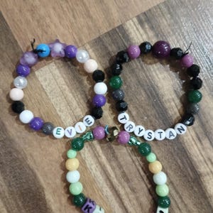 May include: Three beaded bracelets with names spelled out in letter beads. The bracelets feature a variety of colorful beads in shades of purple, green, black, white, and pink. The names "Evie", "Trystan", and "Kingsley" are spelled out.