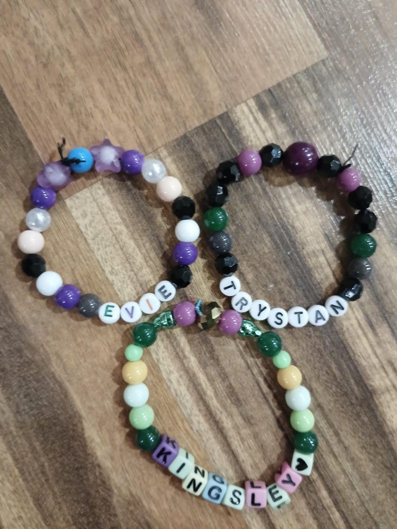 May include: Three beaded bracelets with names spelled out in letter beads. The bracelets feature a variety of colorful beads in shades of purple, green, black, white, and pink. The names "Evie", "Trystan", and "Kingsley" are spelled out.