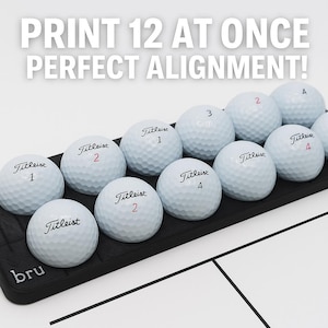 May include: A black tray holds twelve white Titleist golf balls, each with a number and the brand name printed on them. The text "PRINT 12 AT ONCE PERFECT ALIGNMENT!" is displayed above the golf balls.